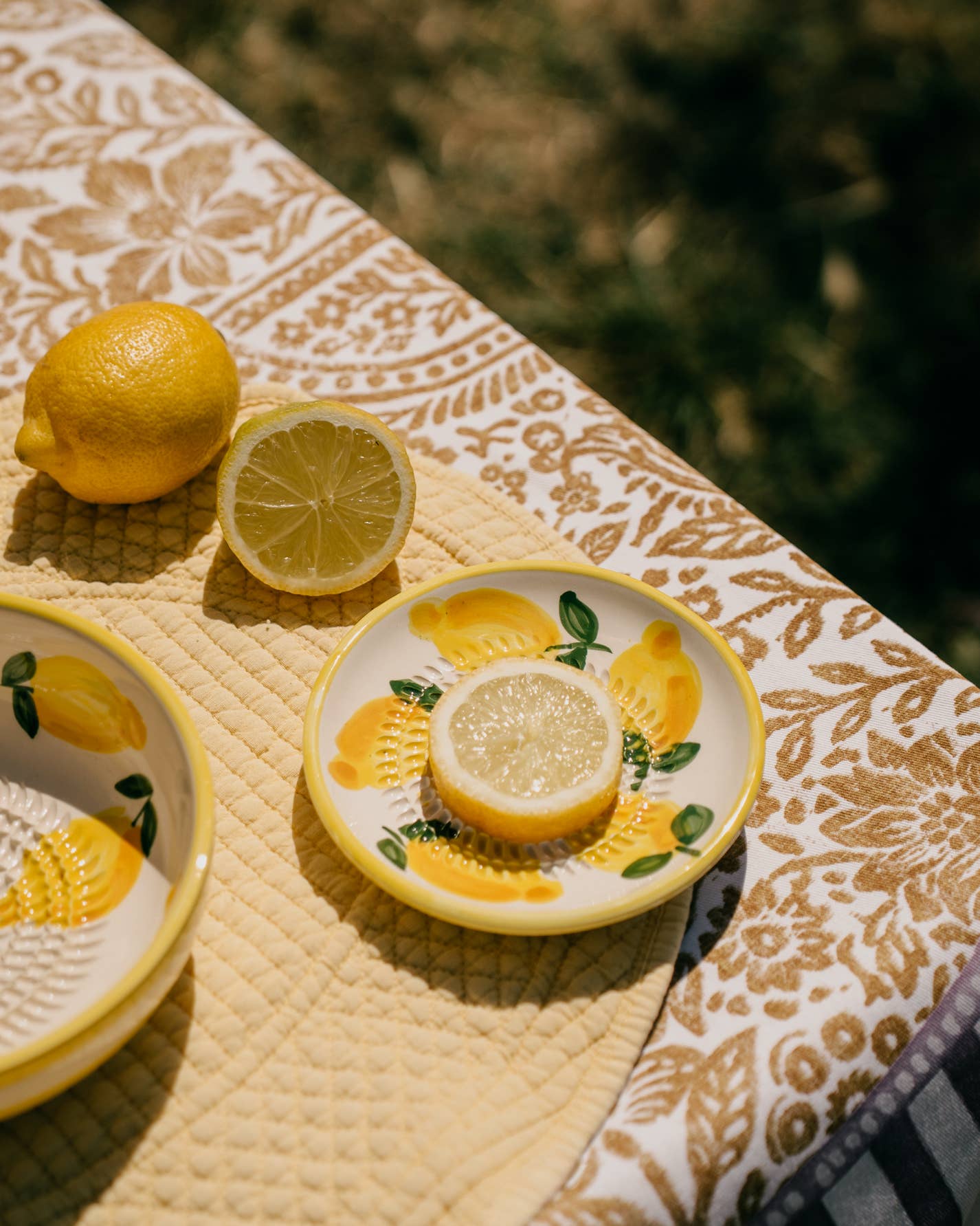 Eine Zitrone, eine halbe Zitrone und eine Zitronenscheibe sitzen auf der Keramik Knoblauchreibe 'Limone' (12,5 x 12,5 x 2,5 cm) von Alfar Tierra Cocida und verleihen einem sonnig gedeckten Tisch mit gelber Steppdecke und Dekotuch einen frischen, sommerlichen Touch.