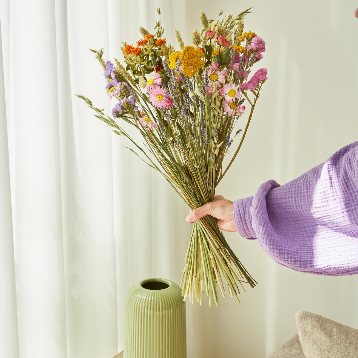 Eine Person mit einem violetten Ärmel hält den Strauß „Wildflowers by Floriette | Trockenblumen ‚Feldblumenstrauß‘ S – M“ neben einer grünen, geriffelten Vase vor einem hellen Hintergrund mit transparenten Vorhängen.