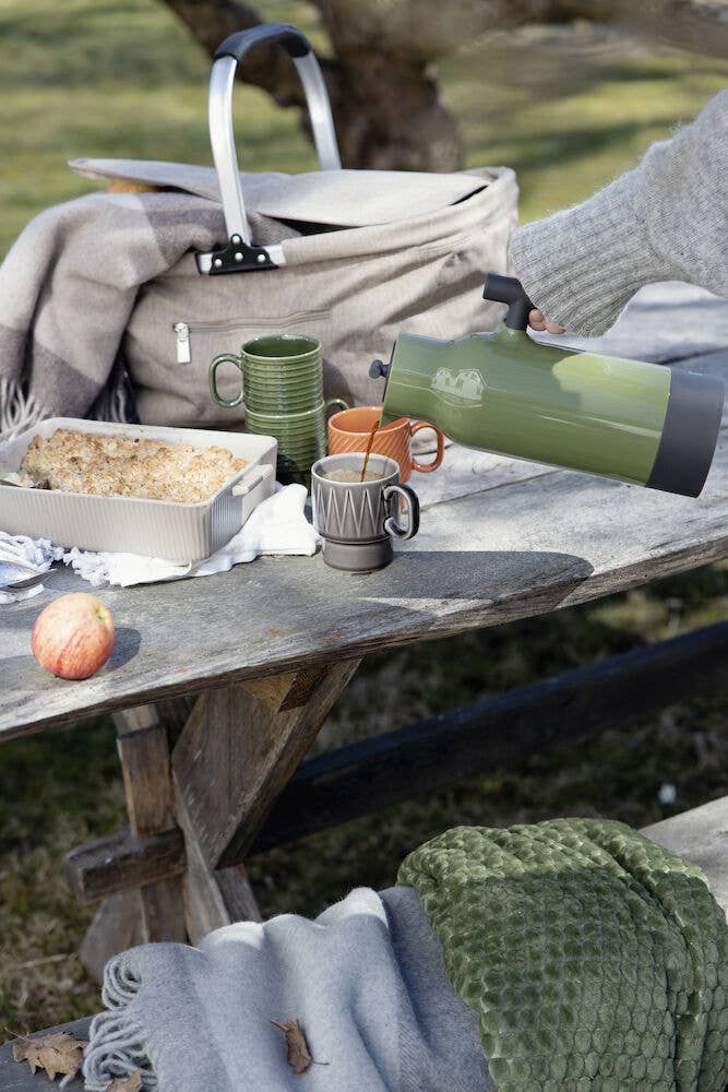 Auf einem rustikalen Picknicktisch mit Decke, Bratpfanne, Apfel und Tasche gießt jemand ein Heißgetränk aus einer grünen Thermoskanne in die Sagaform GmbH Kaffeebecher terrakotta (2-teilig) in einladender Outdoor-Atmosphäre.