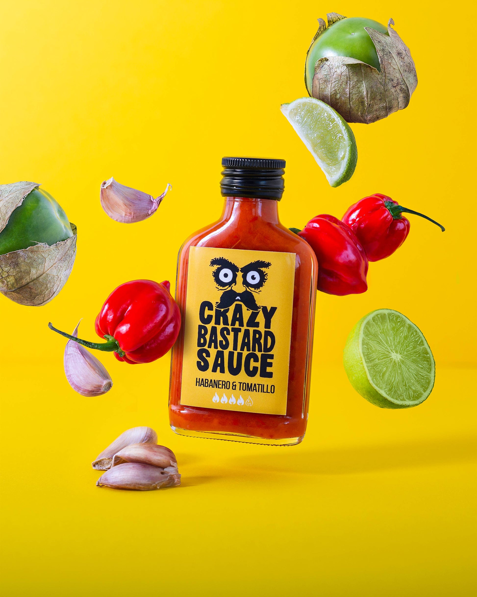 Eine 100-ml-Flasche Crazy Bastard Sauce Habanero & Tomatillo scharfe Soße mit frischen Tomatillos, Habaneros, Knoblauch und Limette auf gelbem Grund - eine kühne Soße für Fans von scharfen Aromen.