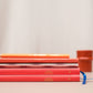 Drei rote Tinne Mia | Notizbücher mit japanischem Verschluss 'Cinnamon' mit goldenen Motiven und blauen Bändern liegen gestapelt auf einer beigen Fläche, darüber ein orangefarbener Bleistift. Eine orangefarbene Tasse steht unscharf im Hintergrund - perfekt für umweltfreundliche Notizen.