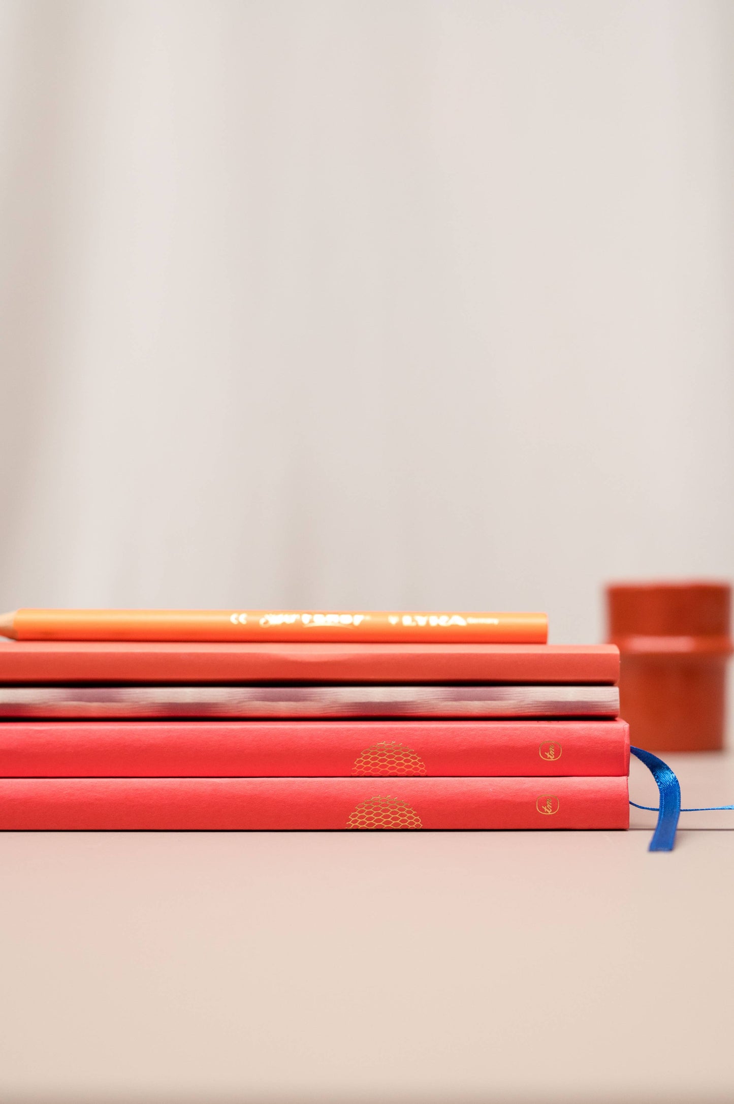 Drei rote Tinne Mia | Notizbücher mit japanischem Verschluss 'Cinnamon' mit goldenen Motiven und blauen Bändern liegen gestapelt auf einer beigen Fläche, darüber ein orangefarbener Bleistift. Eine orangefarbene Tasse steht unscharf im Hintergrund - perfekt für umweltfreundliche Notizen.
