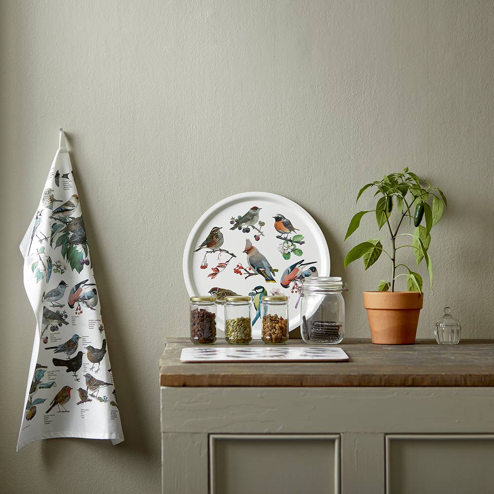 Auf der Küchenarbeitsplatte stehen drei Gläser mit Lebensmitteln, eine Topfpflanze, ein Glasgefäß und ein Teller mit Vogelmotiven. An der Wand hängt das „Koustrupco | Küchentuch mit Gartenvögeln aus 100 % Bio-Baumwolle“ von Koustrupco mit bezaubernden Vogelmotiven. Die schlichte und übersichtliche Anordnung hebt dieses umweltfreundliche Küchentuch als stilvollen Akzent hervor und schafft eine einladende Atmosphäre.