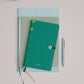 Ein Tinne Mia Notizbuch mit Knopf 'Leinen Smaragd' in teal ist auf zwei anderen gestapelt, ideal als Tagebuch oder Bullet Journal. Ein silberner Stift liegt daneben und eine grüne Tasse sitzt links oben auf einer blassrosa Fläche.