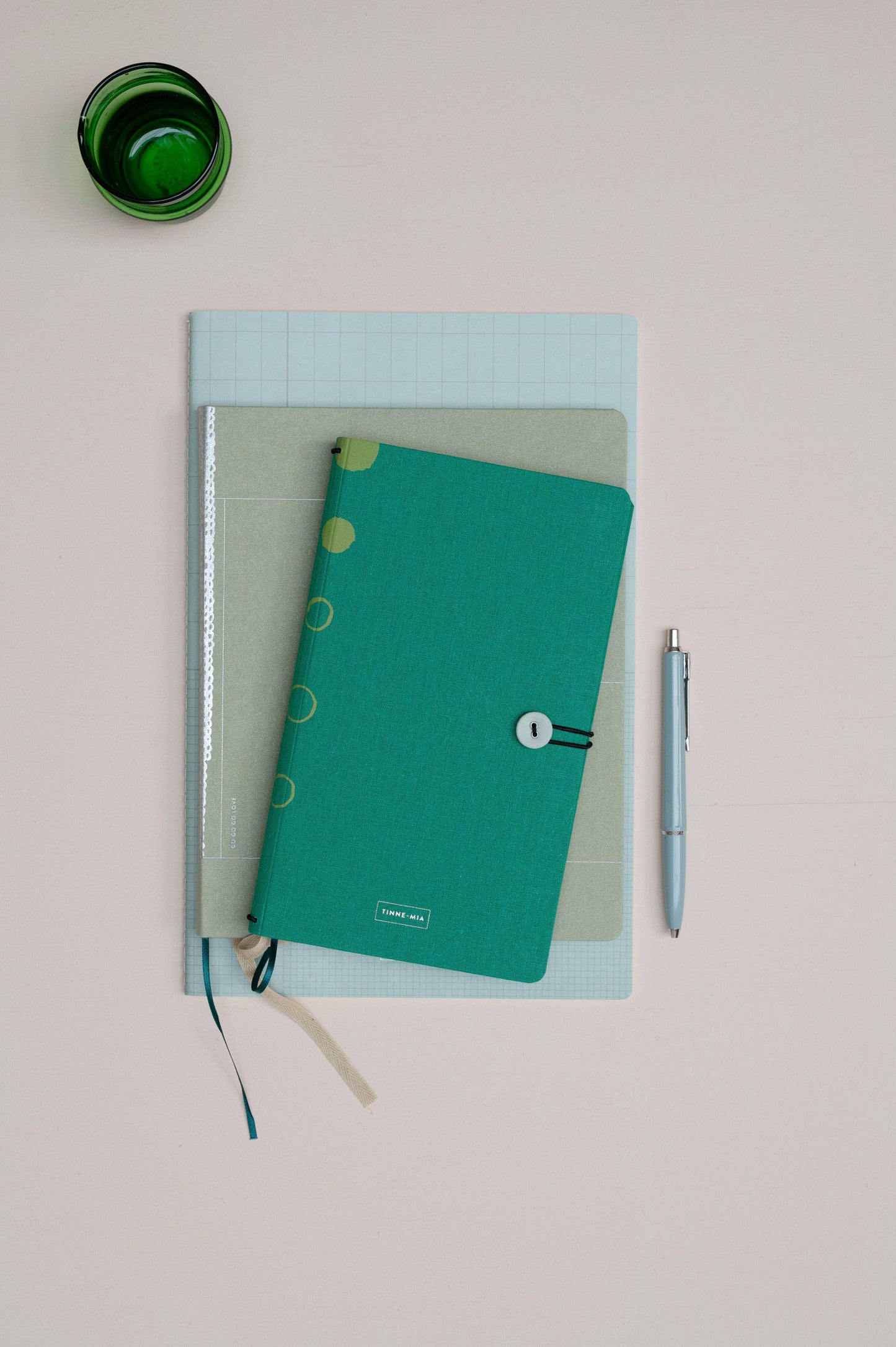 Ein Tinne Mia Notizbuch mit Knopf 'Leinen Smaragd' in teal ist auf zwei anderen gestapelt, ideal als Tagebuch oder Bullet Journal. Ein silberner Stift liegt daneben und eine grüne Tasse sitzt links oben auf einer blassrosa Fläche.