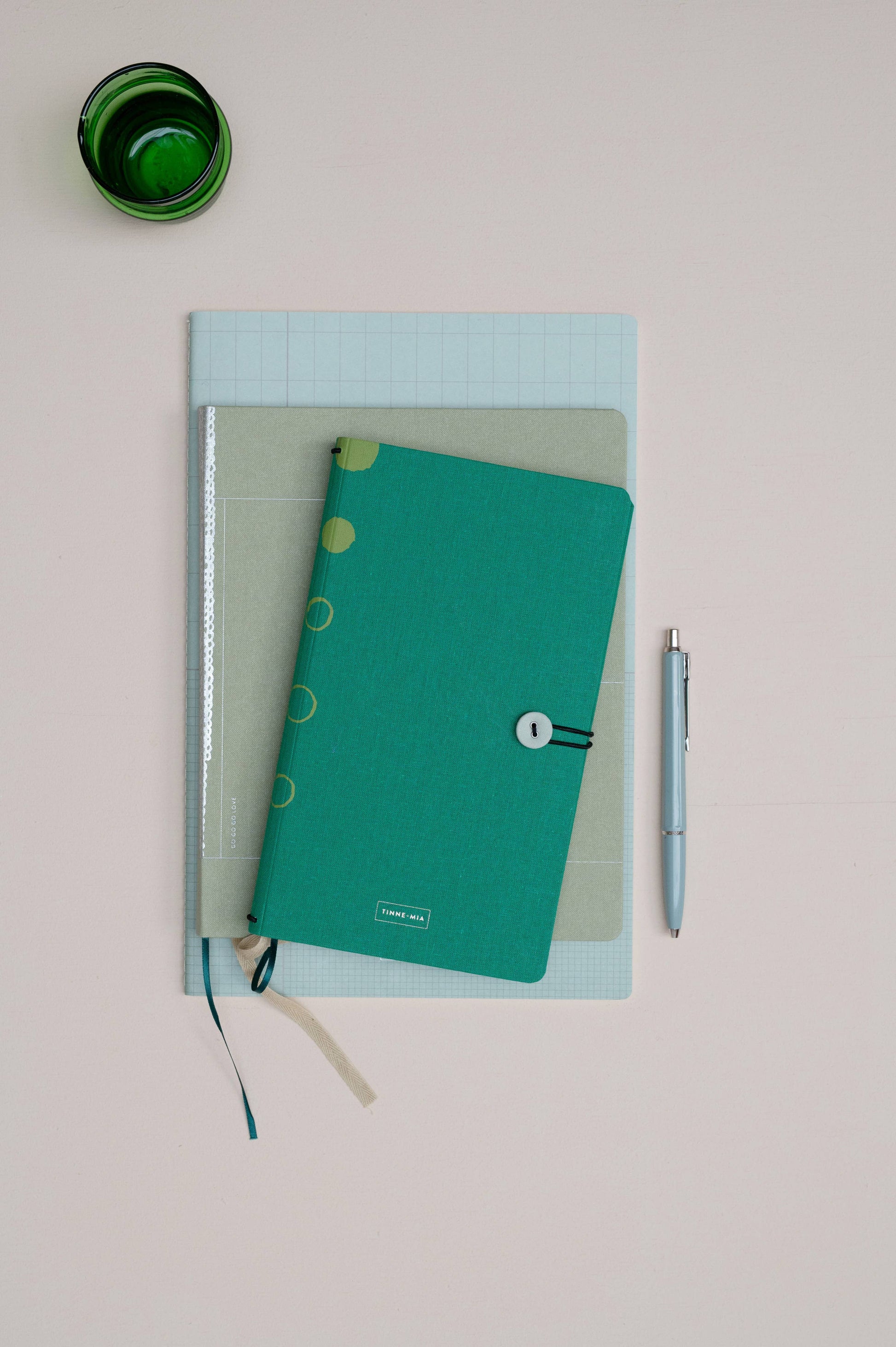 Ein Tinne Mia Notizbuch mit Knopf 'Leinen Smaragd' in teal ist auf zwei anderen gestapelt, ideal als Tagebuch oder Bullet Journal. Ein silberner Stift liegt daneben und eine grüne Tasse sitzt links oben auf einer blassrosa Fläche.