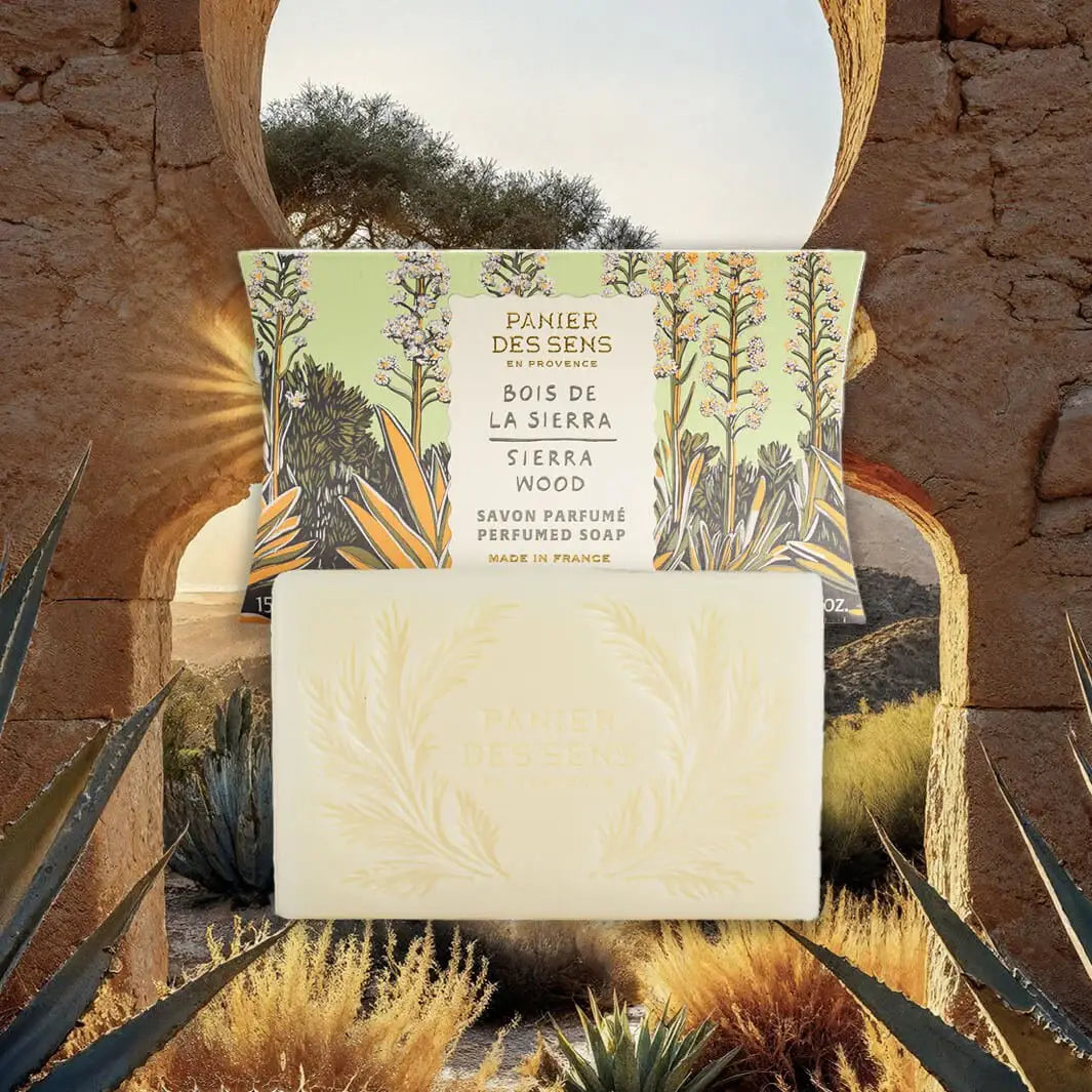 Eine Schachtel Panier Des Sens | Duftseife Sierraholz 150 g und ein graviertes Seifenstück sitzen zwischen Agavenpflanzen an einem Steinbogenfenster mit Blick auf den Sonnenuntergang - und fangen den holzig-würzigen Duft dieser französischen Pflanzenseife ein.
