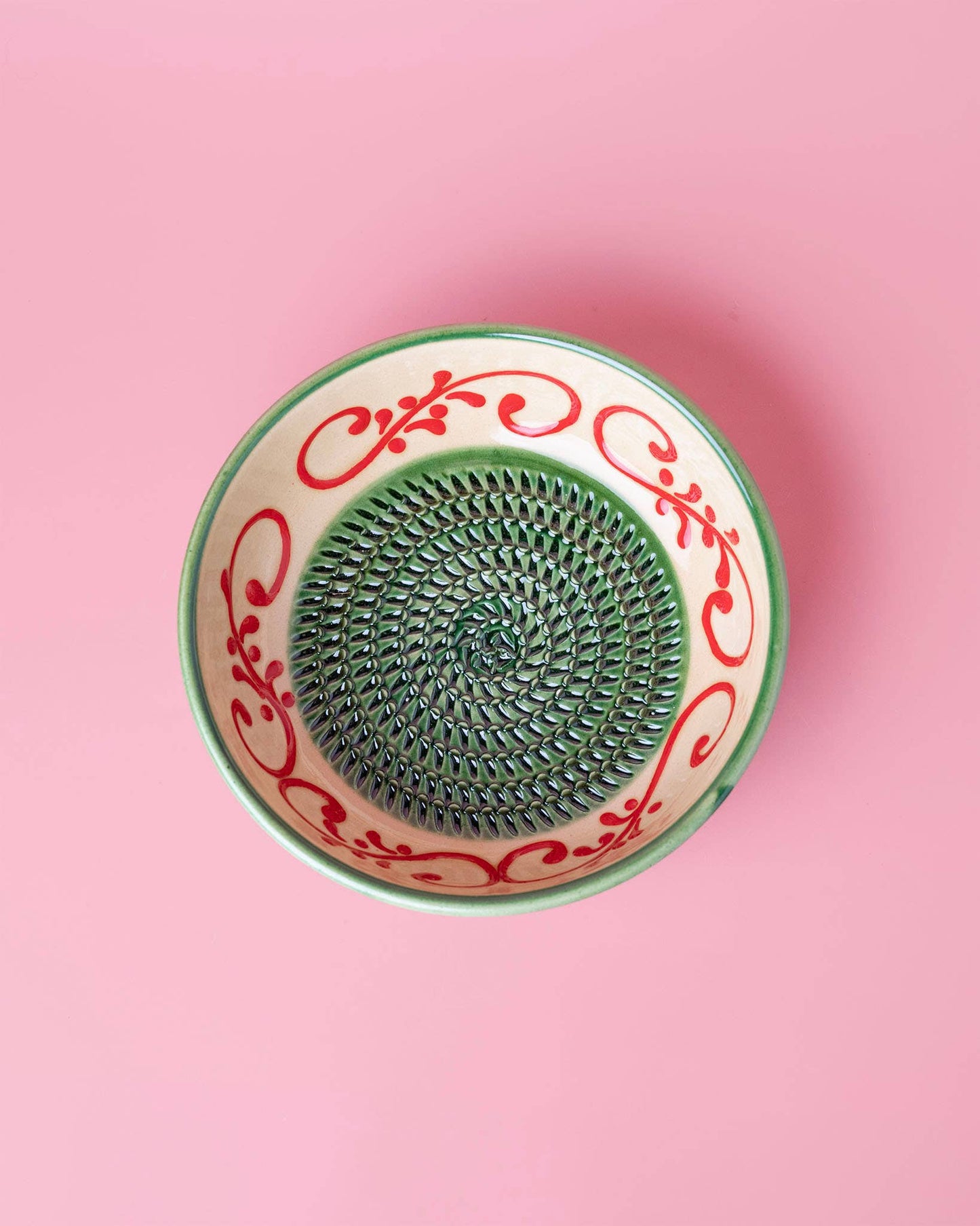 Die Alfar Tierra Cocida 'Alhambra' Knoblauchreibe in Grün (15 x 15 x 5 cm) steht auf einem rosafarbenen Hintergrund und zeigt spanische Handwerkskunst mit einem grünen und roten Blumenmotiv und einem spiralförmigen, strukturierten Zentrum.