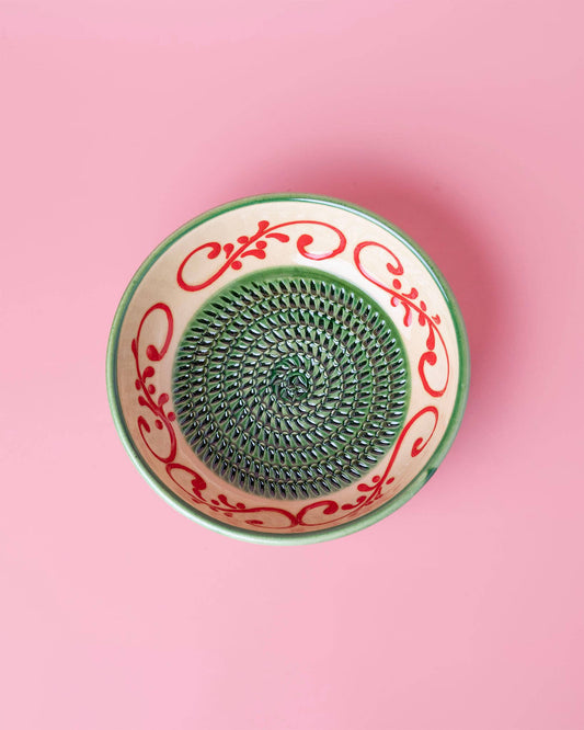 Die Alfar Tierra Cocida 'Alhambra' Knoblauchreibe in Grün (15 x 15 x 5 cm) steht auf einem rosafarbenen Hintergrund und zeigt spanische Handwerkskunst mit einem grünen und roten Blumenmotiv und einem spiralförmigen, strukturierten Zentrum.