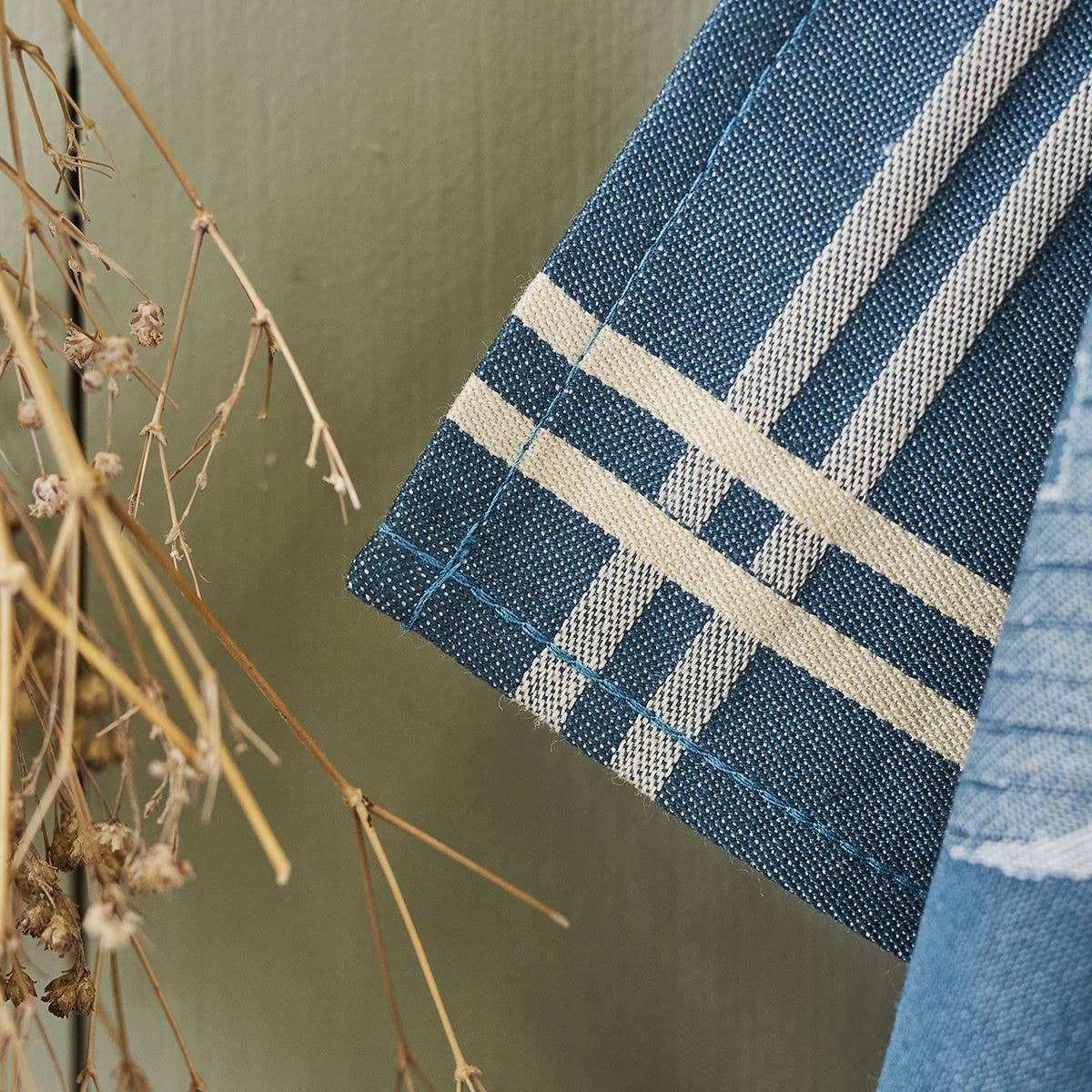 Eine Nahaufnahme des Sylvie Thiriez | Geschirrtuch Jacquard 'Hütten' Blau - mit weißen Karostreifen - hängt neben getrockneten Pflanzenstängeln vor einem gedämpften grünen Hintergrund.