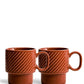 Zwei terrakottafarbene Sagaform Kaffeebecher der Sagaform GmbH, die als 2-teiliges Set verkauft werden, zeichnen sich durch diagonale Strukturmuster, große Griffe und ein modernes geometrisches Design aus. Sie sind nebeneinander auf einem weißen Hintergrund abgebildet.