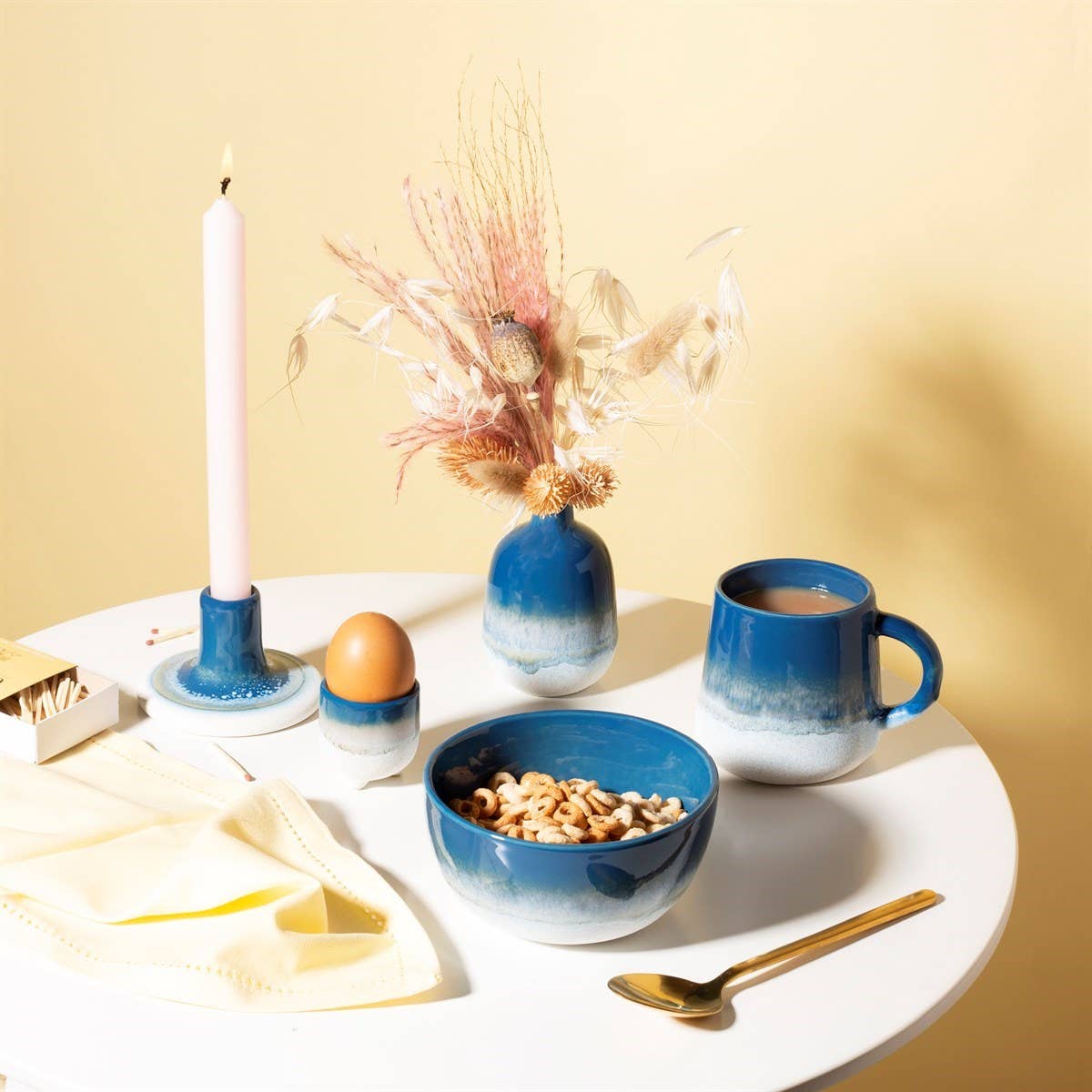 Auf einem weißen Tisch stehen eine blaue Tasse, eine blaue Müslischale in einem ozeanblauen Farbton, ein Ei in einem Halter, eine Kerze und die blaue Vase „Mojave Glaze“ von Sass & Belle Europe (12,5 cm), gefüllt mit Trockenblumen für einen dezenten Boho-Charme, sowie eine Serviette, einem goldenen Löffel und einer Streichholzschachtel – alles vor einem blassgelben Hintergrund.