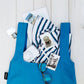 Eine Notabag | „Original Blue“ von Notabag liegt auf weißem Holz, gefüllt mit Kleidung, Sonnenbrille, Fotos, Postkarte, Gerät und Stift – die vielseitige Tasche ist Rucksack und Handtasche in einem.