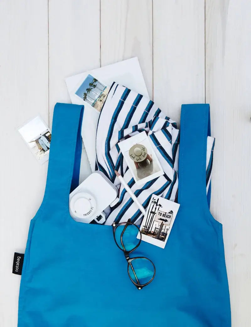 Eine Notabag | „Original Blue“ von Notabag liegt auf weißem Holz, gefüllt mit Kleidung, Sonnenbrille, Fotos, Postkarte, Gerät und Stift – die vielseitige Tasche ist Rucksack und Handtasche in einem.