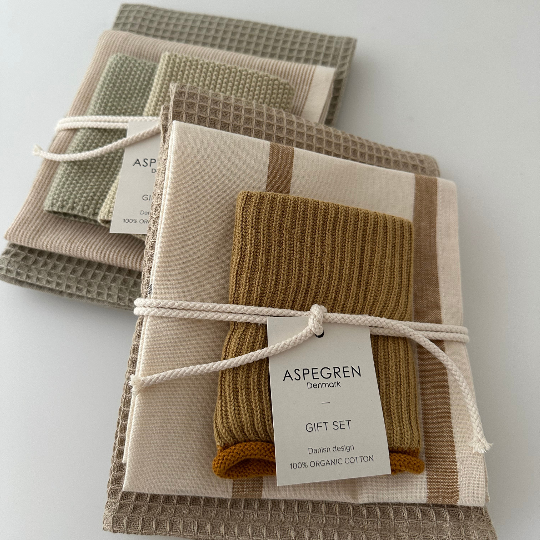 Zwei gefaltete Aspegren Denmark Küchentuch-3er-Geschenksets in Naturtönen sind mit einer Schnur zusammengebunden. Jedes Set enthält drei Geschirrtücher in neutralen, erdigen Farbtönen. Auf dem Etikett steht „Aspegren Denmark Geschenkset, dänisches Design, 100 % Bio-Baumwolle“. Diese stilvollen Küchentextilien verbinden Nachhaltigkeit mit skandinavischer Eleganz – perfekt für jedes moderne Zuhause.