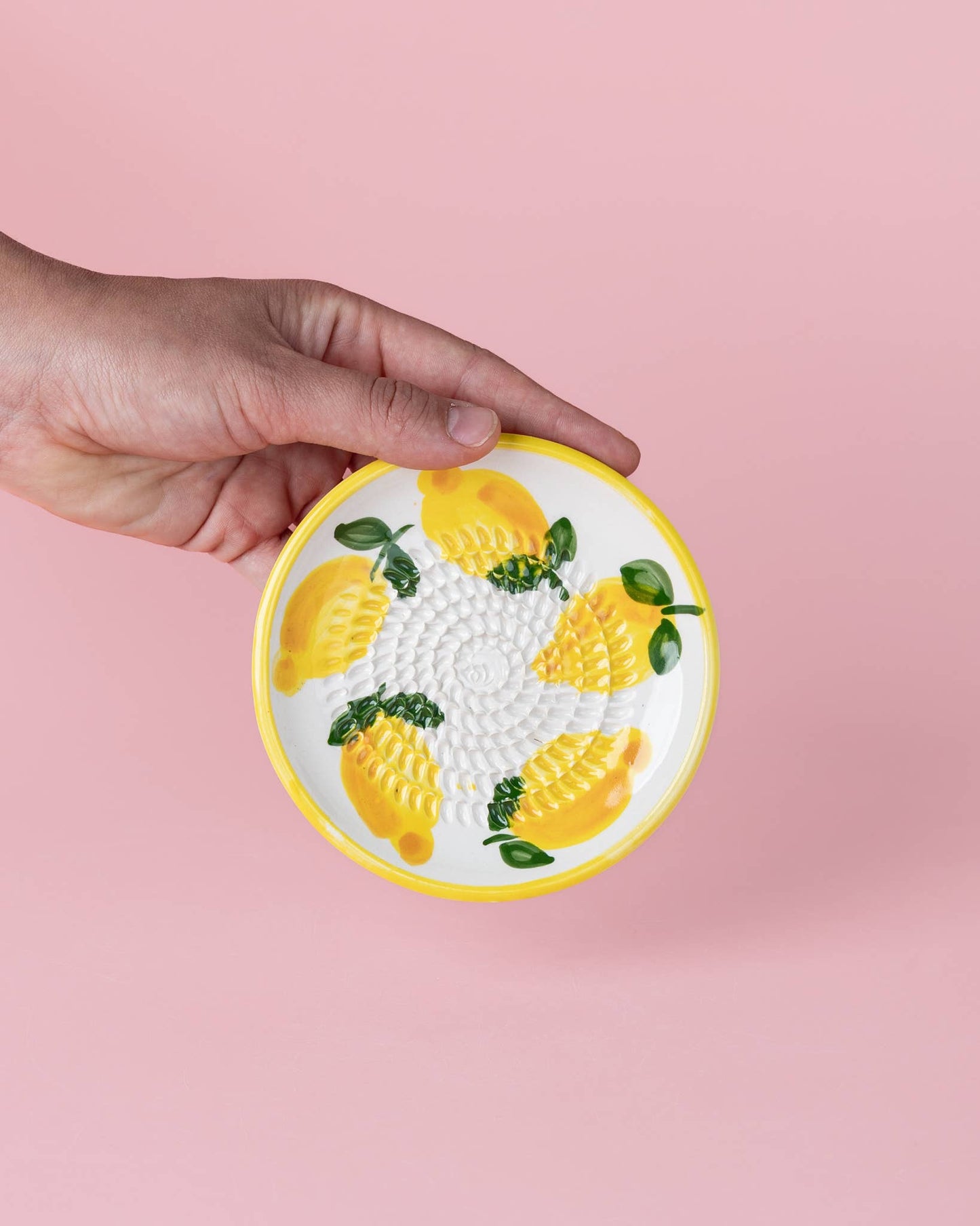 Eine Hand hält die Alfar Tierra Cocida 'Limone' Keramik Knoblauchreibe (12,5 x 12,5 x 2,5 cm), einen kleinen, runden Keramikteller mit gelbem Rand und gemaltem Zitronenmotiv vor einem hellrosa Hintergrund.