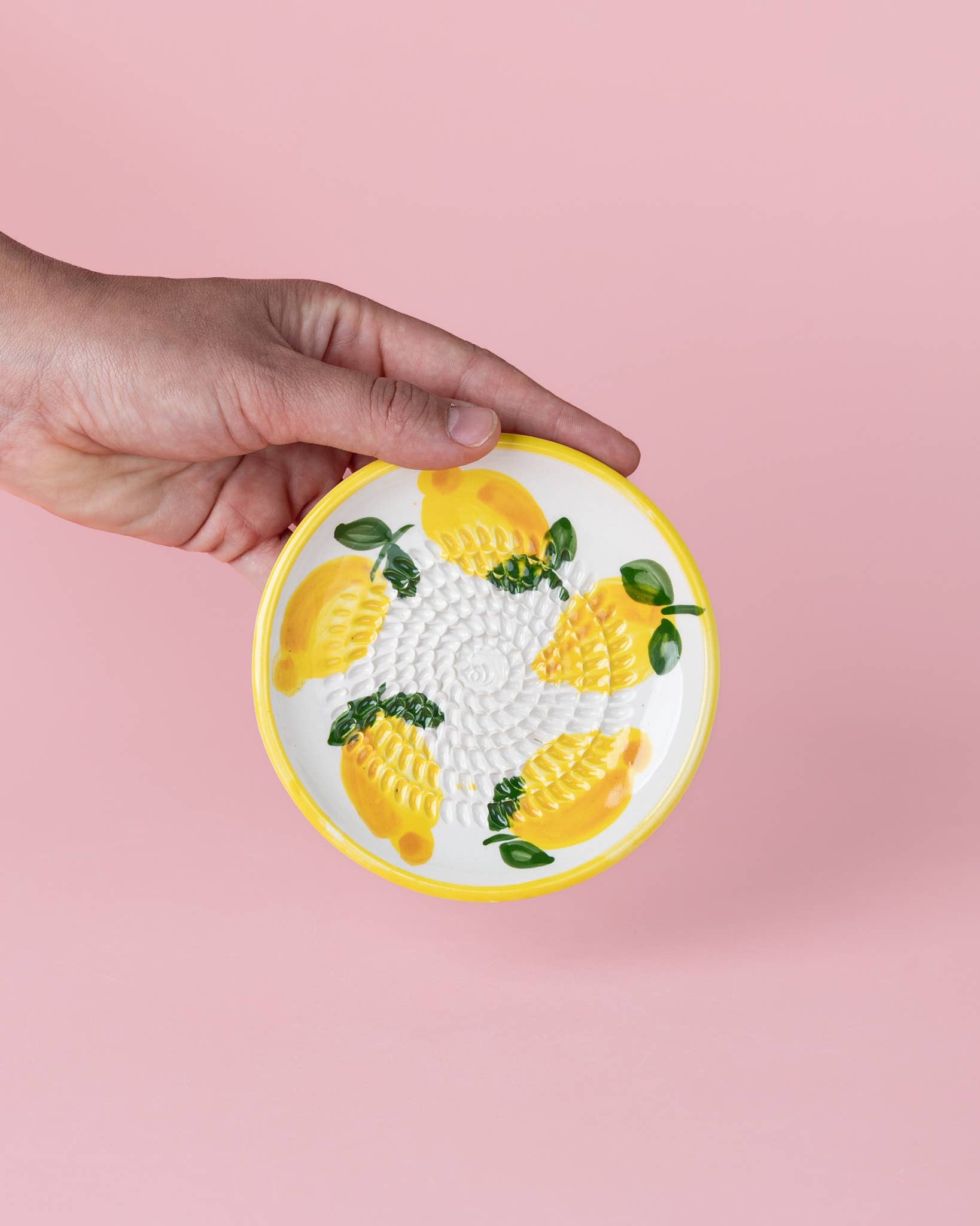 Eine Hand hält die Alfar Tierra Cocida 'Limone' Keramik Knoblauchreibe (12,5 x 12,5 x 2,5 cm), einen kleinen, runden Keramikteller mit gelbem Rand und gemaltem Zitronenmotiv vor einem hellrosa Hintergrund.
