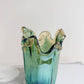 Die mittelgroße Vase „Venezia“ von Murano Dream aus grünem Murano-Glas (H 25 cm, Ø 14 cm) mit gewelltem Rand und gelben Akzenten besticht vor schlichtem Hintergrund durch ihre edle, künstlerische Ausstrahlung.