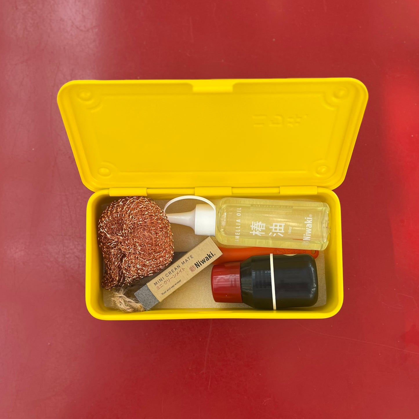 Eine gelbe Niwaki 'Mini Clean Mate' Box auf roter Oberfläche enthält einen Kupferschrubber, Rost- und Harzentferner mit Pumpe, Seife in braunem Papier und einen schwarzen Behälter - ideal für die Reinigung von Gartenwerkzeugen.