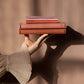 Eine Hand in einem durchsichtigen, plissierten Ärmel hält zwei FSC-zertifizierte Bücher, auf denen das Notizbuch mit Knopf 'Leinen Rose Dawn' von Tinne Mia gestapelt ist, vor einem sanft beleuchteten beigen Hintergrund mit sanften Schatten.