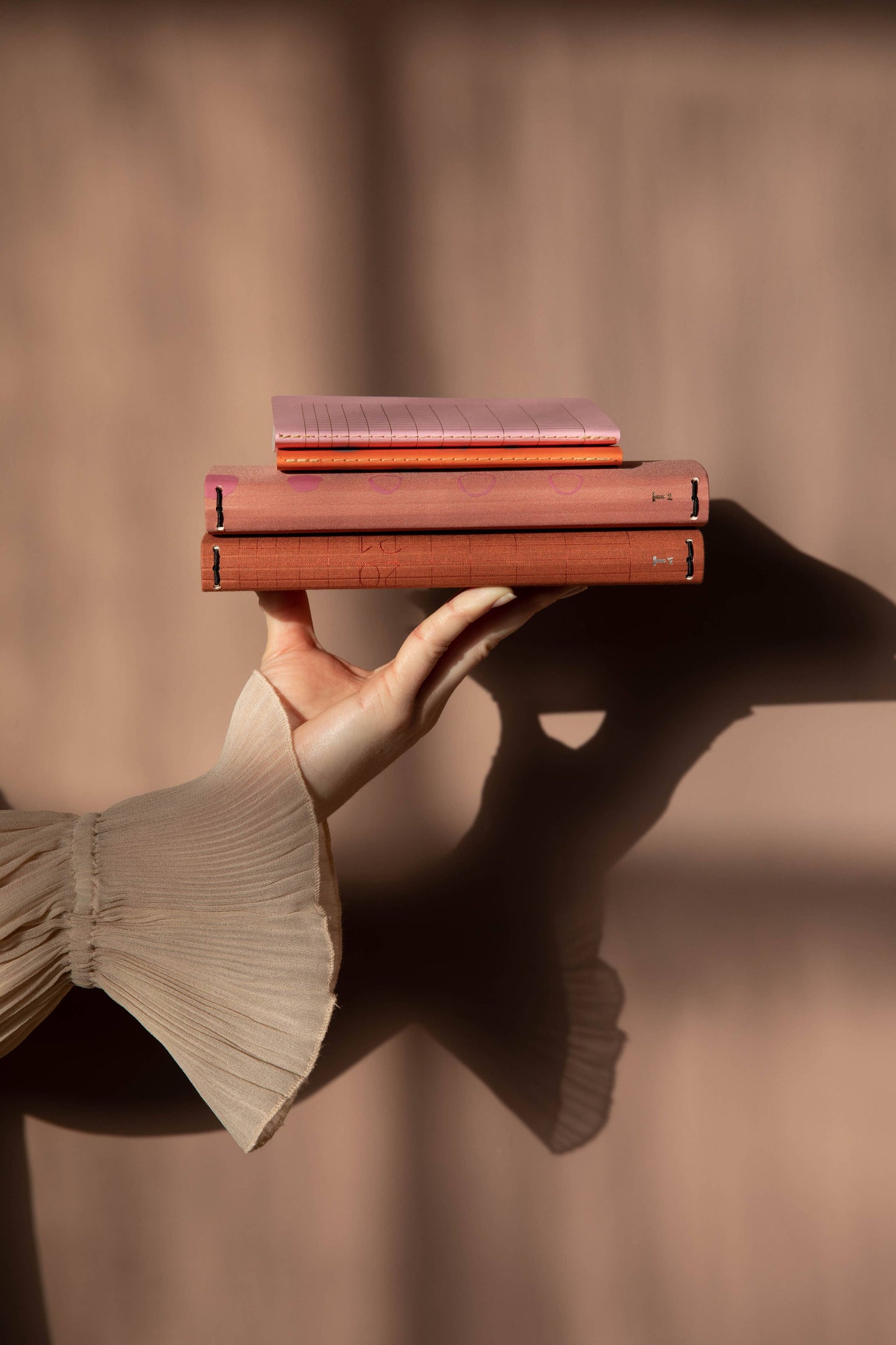Eine Hand in einem durchsichtigen, plissierten Ärmel hält zwei FSC-zertifizierte Bücher, auf denen das Notizbuch mit Knopf 'Leinen Rose Dawn' von Tinne Mia gestapelt ist, vor einem sanft beleuchteten beigen Hintergrund mit sanften Schatten.
