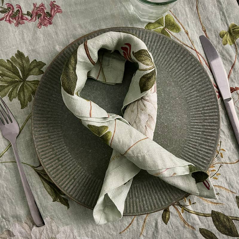 Auf einem grauen, strukturierten Teller liegt die Leinenserviette „Flora Danica Atlas“ von Koustrupco (45 × 45 cm), die wie ein gedrehtes Band gefaltet ist, auf einer passenden Tischdecke mit Blumenmuster, dazu Besteck und ein Glas.