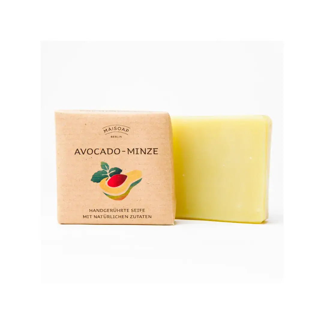 Ein gelber Seifenriegel von Maisoap | Avocado-Minze-Seife (90 g) steht neben seiner braunen Papierverpackung, die mit einer Abbildung einer Avocado und Minzblättern bedruckt ist. Der Text beschreibt sie als eine natürlich hergestellte, handgesiedete Seife aus Berlin von Maisoap, die aus natürlichen Inhaltsstoffen hergestellt wird.