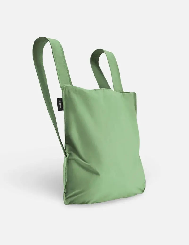 Eine hellolivgrüne Notabag | „Olive“ von Notabag – Rucksack und Tasche in einem – mit zwei Schulterriemen steht aufrecht vor einem schlichten, hellen Hintergrund.
