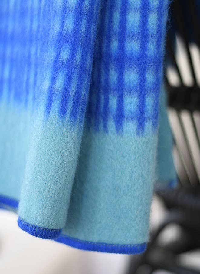 Eine Nahaufnahme der Blauen Tagesdecke "Darling" aus Lammwolle von Sofie Sjöström Design. Sie zeigt den gemütlichen, strukturierten Stoff mit einem blauen Verlaufsmuster, karierten Linien auf der Oberseite und einem einfarbigen hellblauen Rand auf der Unterseite.