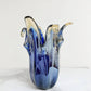 Die Murano Dream „Giusy“-Vase aus blauem Murano-Glas (H 24, Ø 19 cm) vereint italienische Glaskunst mit fließenden, gewellten Rändern. Präsentiert auf weißem Marmorboden vor schlichtem Hintergrund.