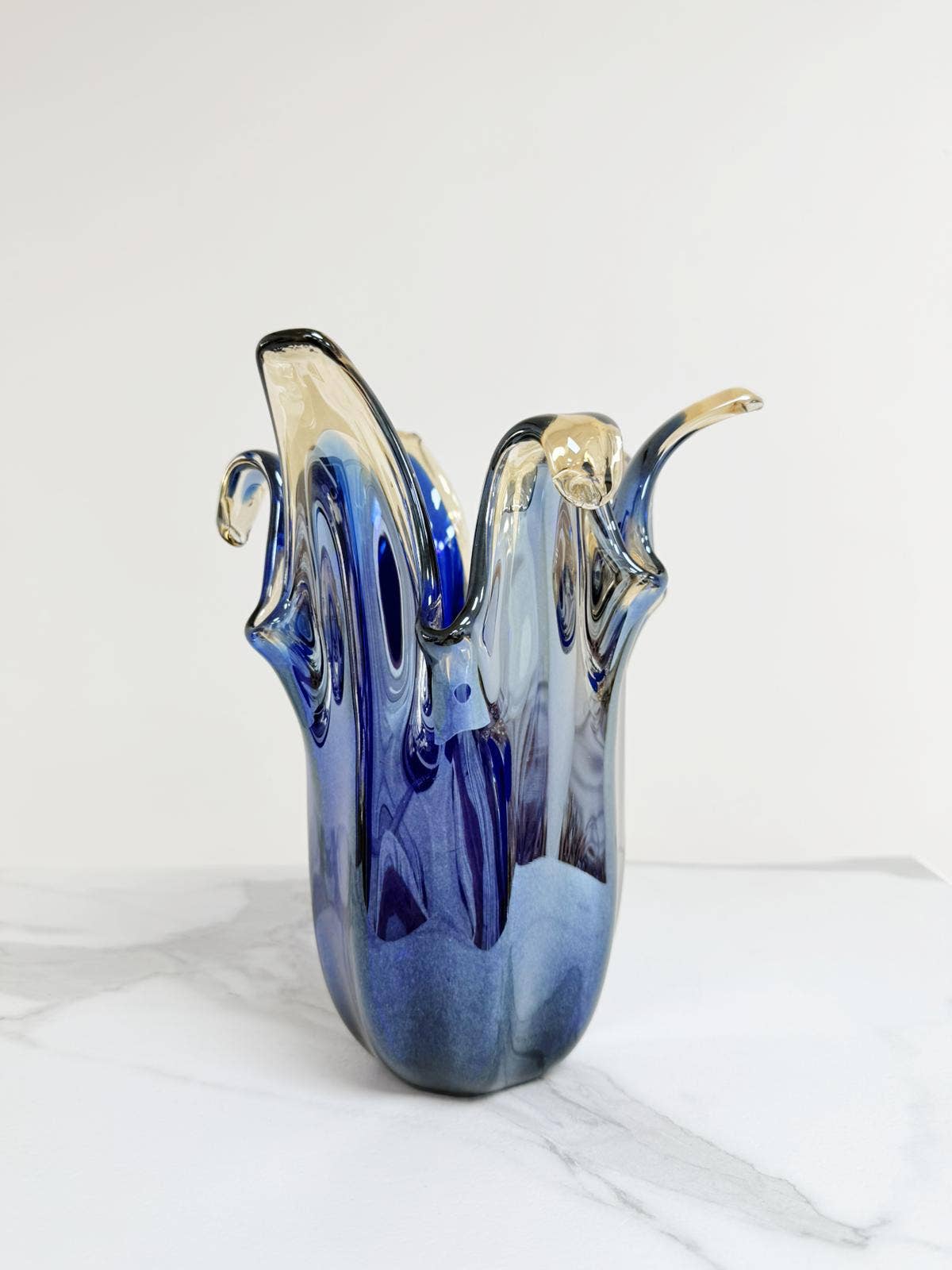 Die Murano Dream „Giusy“-Vase aus blauem Murano-Glas (H 24, Ø 19 cm) vereint italienische Glaskunst mit fließenden, gewellten Rändern. Präsentiert auf weißem Marmorboden vor schlichtem Hintergrund.