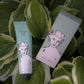 Eine Tube und eine Schachtel der "Aloe Vera"-Handcreme von Archivist Gallery mit einem illustrierten Frauengesicht ruhen auf üppigen grünen Blättern, die die natürliche Schönheit dieser veganen Handcreme unterstreichen.