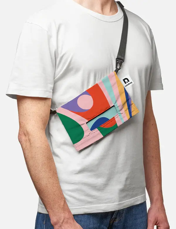 Eine Person trägt ein schlichtes weißes T-Shirt und eine Notabag | 'Flow' Leichte Umhängetasche von Notabag mit einem farbenfrohen abstrakten geometrischen Muster in Grün, Rosa, Lila, Blau, Orange und Rot und einem schwarzen verstellbaren Riemen.