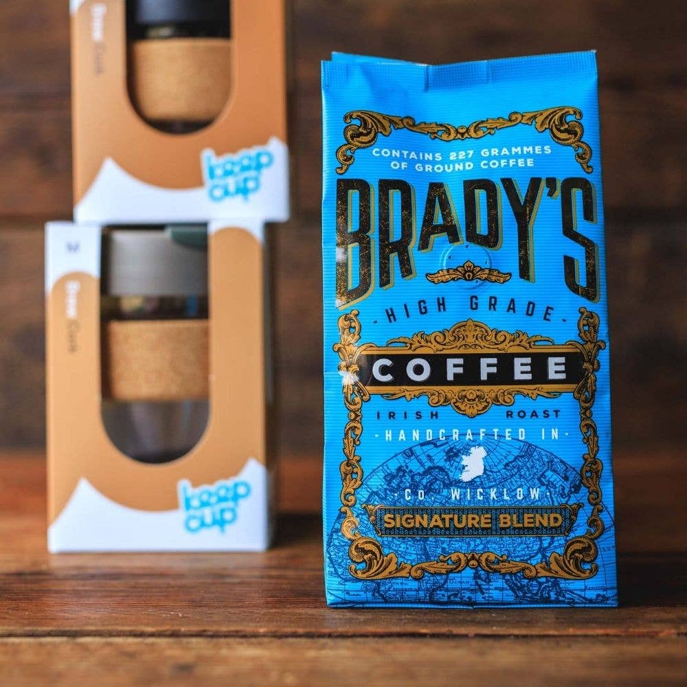 Ein blauer 227-g-Beutel gemahlener Kaffee der Brady's Coffee Company Signature Blend steht vor zwei verpackten KeepCup-Mehrwegbechern auf einer hölzernen Unterlage, deren goldene und weiße Details die Premium-Qualität unterstreichen.