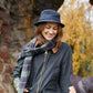 Eine Frau lächelt im Freien und trägt den Wachstropfen Krempenhut von Tweedmill Textiles, einen karierten Schal und eine grüne Jacke. Im Hintergrund sind Herbstblätter und eine Steinmauer zu sehen, während sie nach unten schaut.