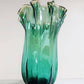 Die „Murano Dream Große ‚Fazzoletto‘-Vase“ aus grünem Murano-Glas (H 32 Ø 24 cm) zeichnet sich durch hohe, wellenförmige Ränder mit einer geriffelten Struktur und einem Farbverlauf in Grüntönen aus, die sich von einem schlichten, hellen Hintergrund abheben.