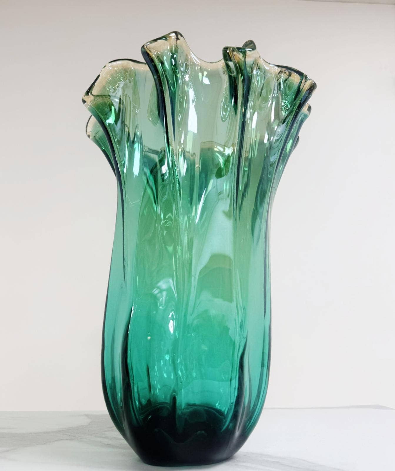 Die „Murano Dream Große ‚Fazzoletto‘-Vase“ aus grünem Murano-Glas (H 32 Ø 24 cm) zeichnet sich durch hohe, wellenförmige Ränder mit einer geriffelten Struktur und einem Farbverlauf in Grüntönen aus, die sich von einem schlichten, hellen Hintergrund abheben.