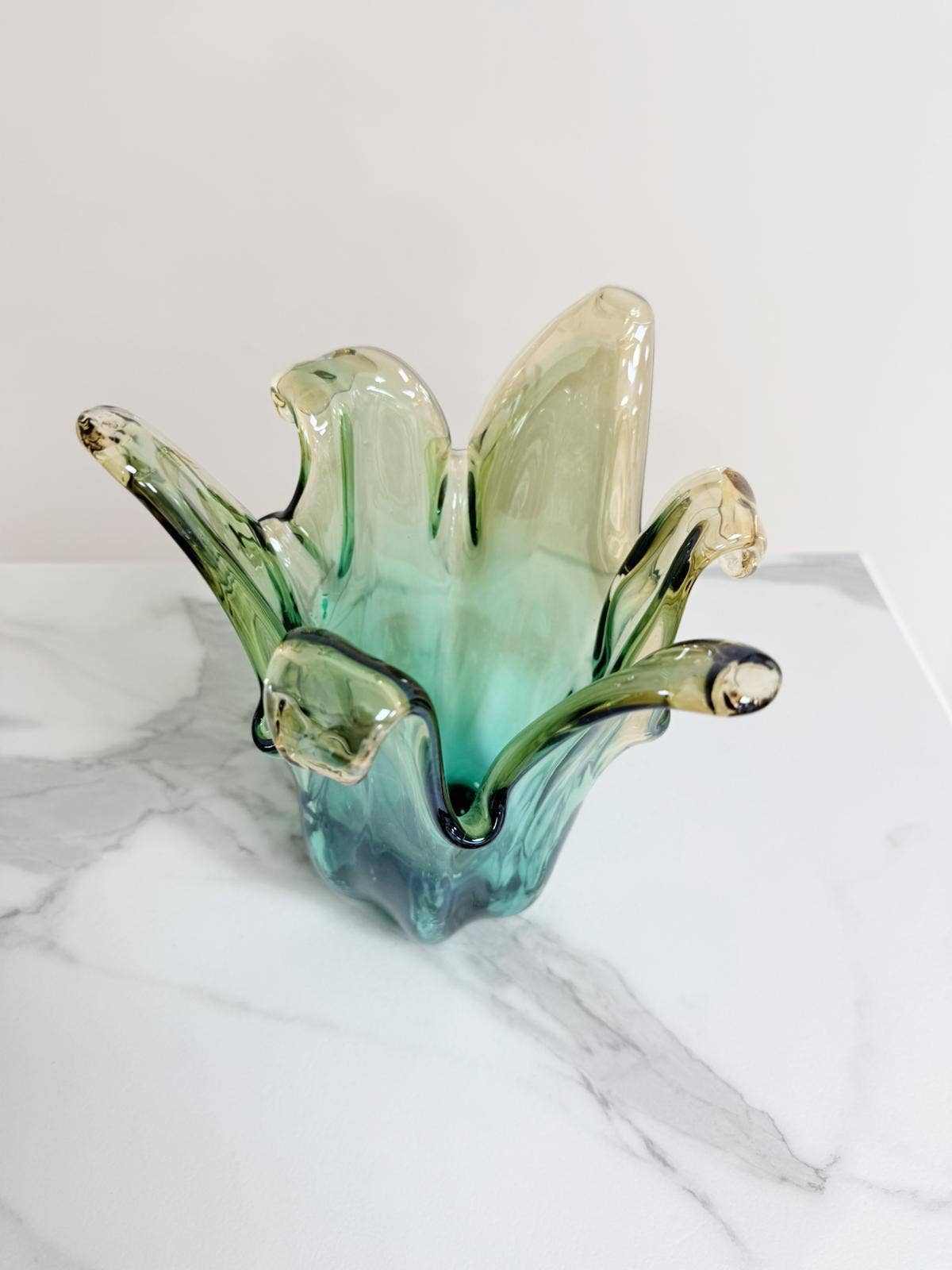 Die mittelgroße „Giusy“-Vase aus grünem Murano-Glas (H 24, Ø 18 cm) der Marke Murano Dream besticht durch handgefertigte, wellenförmig geschwungene Ränder in Grün- und Beigetönen und steht auf weißem Marmor vor hellem Hintergrund.