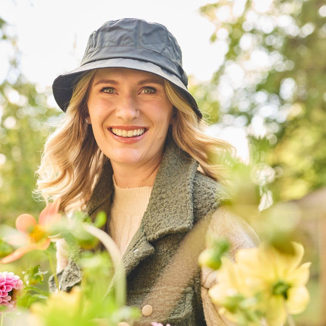 Eine Frau, die den „Wachstropfen“-Krempenhut von Tweedmill Textiles in Schwarz trägt, lächelt im Freien, umgeben von blühenden gelben und rosa Blumen, während im Hintergrund Sonnenlicht durch die Bäume scheint. Der in Großbritannien hergestellte Hut verleiht ihrem Look inmitten der farbenfrohen Gartenszene einen Hauch von britischem Charme.