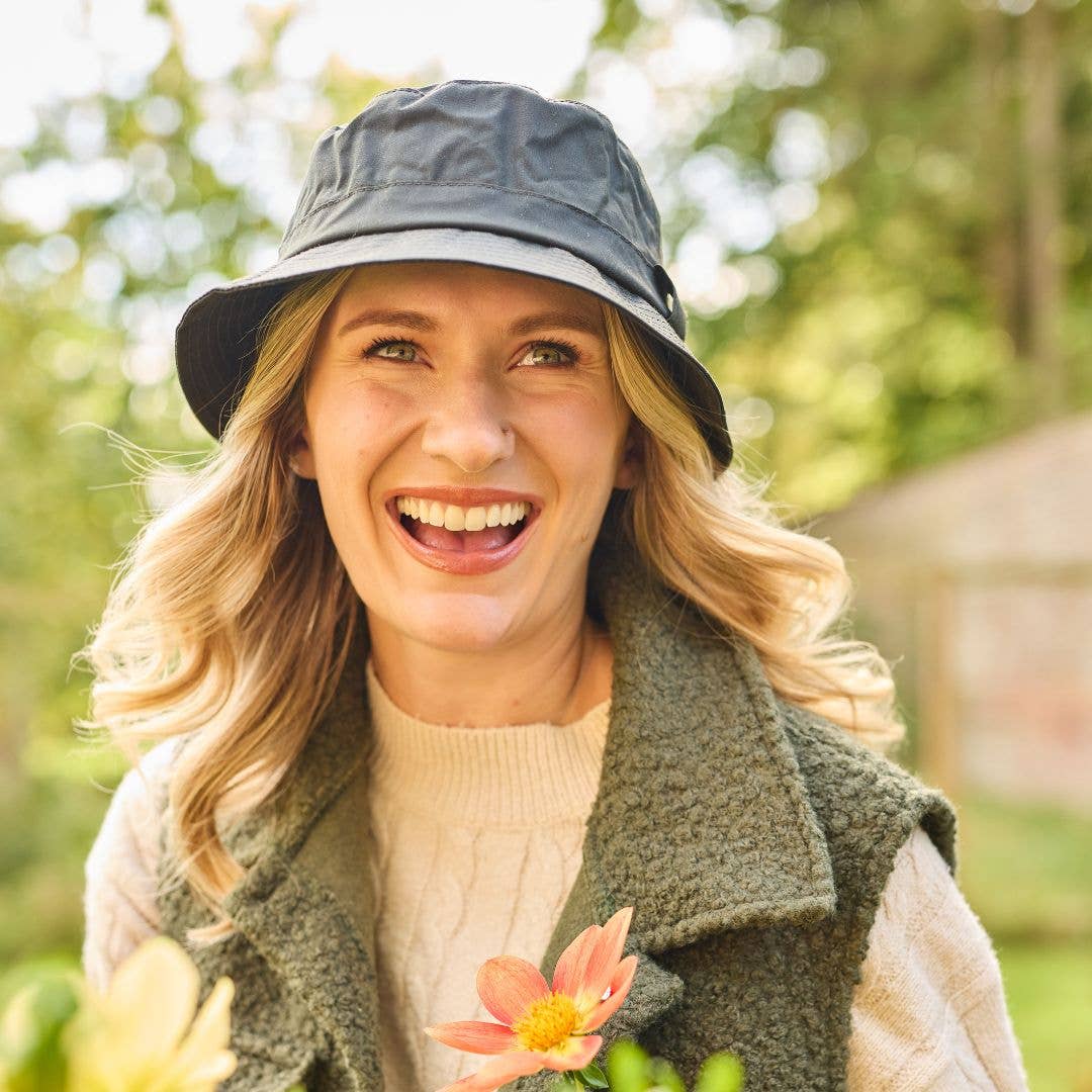 Eine lächelnde Frau mit blonden Haaren trägt den „Wachstropfen“-Krempenhut aus gewachster Baumwolle von Tweedmill Textiles mit breiter Krempe sowie eine grüne Weste und einen cremefarbenen Pullover, während sie im Freien steht und Blumen in der Hand hält. Der stilvolle schwarze Bucket-Hat wird in Großbritannien hergestellt. Im Hintergrund sind Bäume und eine Mauer sanft verschwommen.
