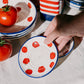 Eine Hand hält die Alfar Tierra Cocida | Kleine Keramik-Knoblauchreibe „Pomodoro“, handgefertigt in Spanien, die mit roten Tomatenmotiven verziert ist und von weiterer traditioneller Keramik von Alfar Tierra Cocida, frischen Tomaten und gestreiften Behältern auf einem weißen Tuch umgeben ist.