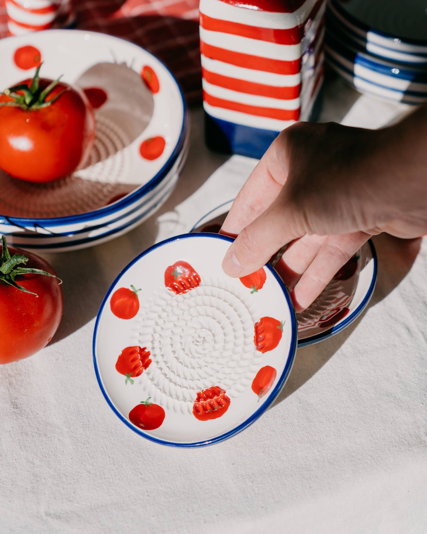 Eine Hand hält die Alfar Tierra Cocida | Kleine Keramik-Knoblauchreibe „Pomodoro“, handgefertigt in Spanien, die mit roten Tomatenmotiven verziert ist und von weiterer traditioneller Keramik von Alfar Tierra Cocida, frischen Tomaten und gestreiften Behältern auf einem weißen Tuch umgeben ist.
