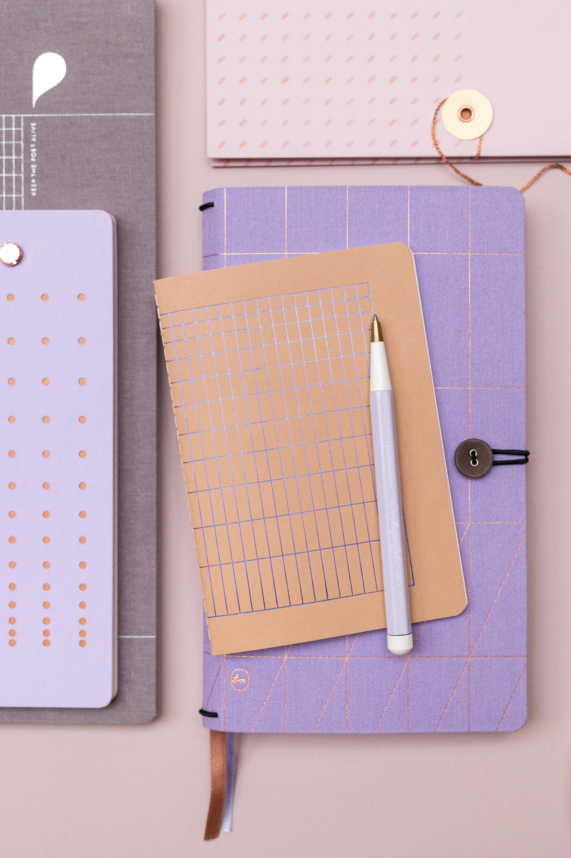 Das Tinne Mia Notizbuch A6 'Nougat Grid' mit geometrischen Mustern, ein brauner Notizblock mit gepunktetem Raster und ein weißer Stift sind ordentlich auf einer hellvioletten Oberfläche angeordnet.