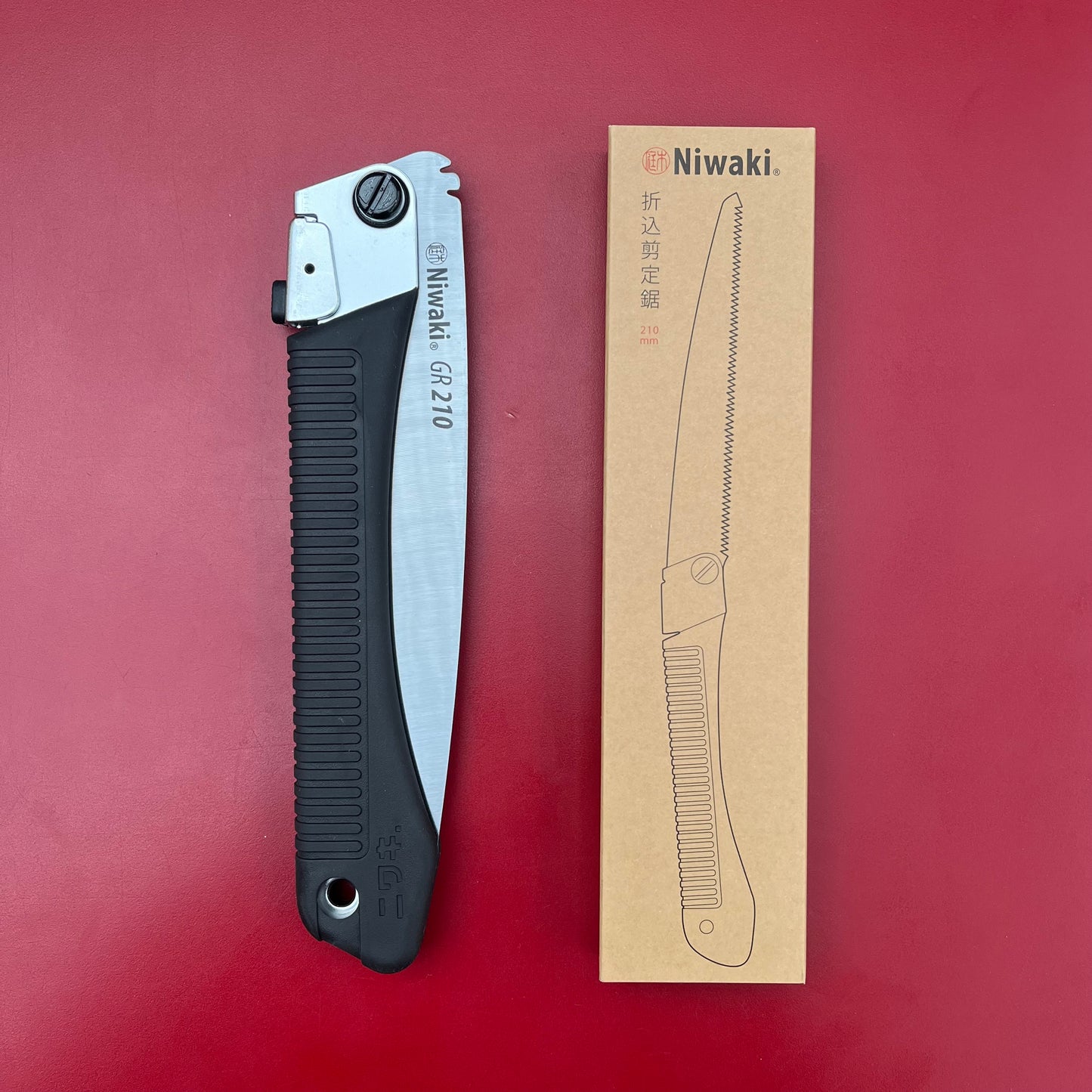 Eine Niwaki 'GR 210 Folding Saw' Klappsäge mit schwarzem Griff und silbernem Sägeblatt wird neben ihrer braunen Schachtel gezeigt, die eine Outdoor Säge Strichzeichnung zeigt, alles vor einem kräftigen roten Hintergrund.