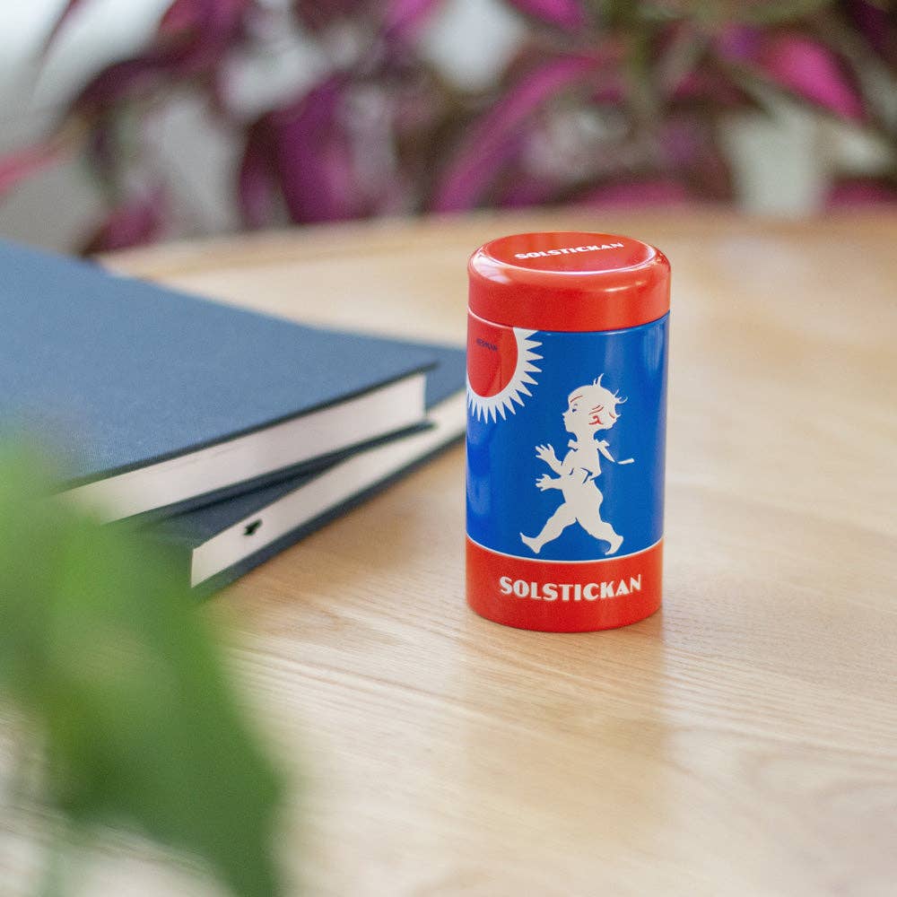 Eine Solstickan Design | Streichholzdose Originaldesign in Blau und Rot mit weißer Jungensilhouette von Solstickan Design steht auf einem Holztisch neben einem dunklen Notizbuch, grünen und lilafarbenen Pflanzen im Hintergrund.
