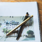 Ein schwarzer Druckbleistift und die ENTREPÔT YSTUDIO Klassischer Revolve Kugelschreiber Feder (Messing) sind auf einem aufgeschlagenen Buch, das Fotos eines mit Pflanzen gefüllten, sonnenbeschienenen Raums zeigt, zu einem "X" gekreuzt.