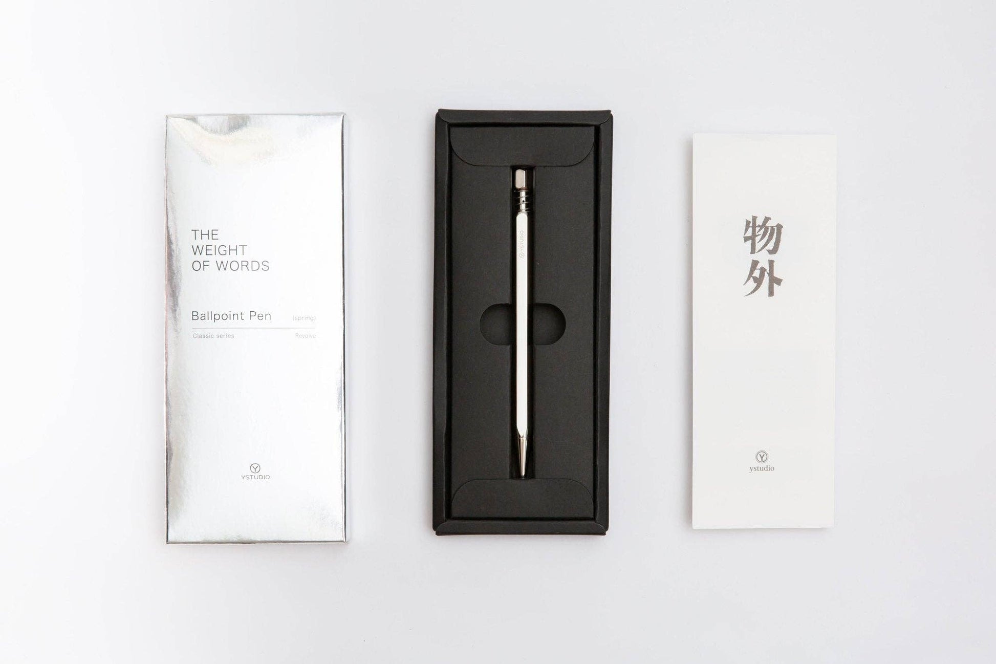 Ein Klassischer Drehkugelschreiber Feder (Glänzendes Silber) von ENTREPÔT, in einer schwarzen Geschenkbox, sitzt zwischen einer weißen THE WEIGHT OF WORDS Kugelschreiber-Verpackung und einer weißen Karte mit chinesischen Schriftzeichen auf weißem Hintergrund.