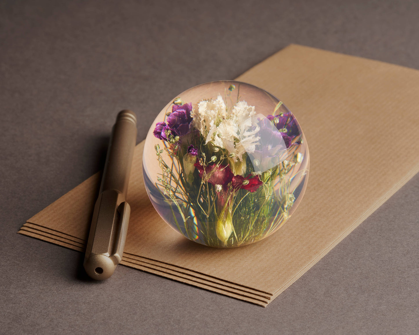 Ein goldener Stift und braune Briefumschläge liegen auf einer grauen Fläche. Darüber befindet sich der Hafod Grange | Briefbeschwerer mit gemischter Flora von Hafod Grange, der bunte Trockenblumen zeigt - ein charmantes Schreibtisch-Accessoire.