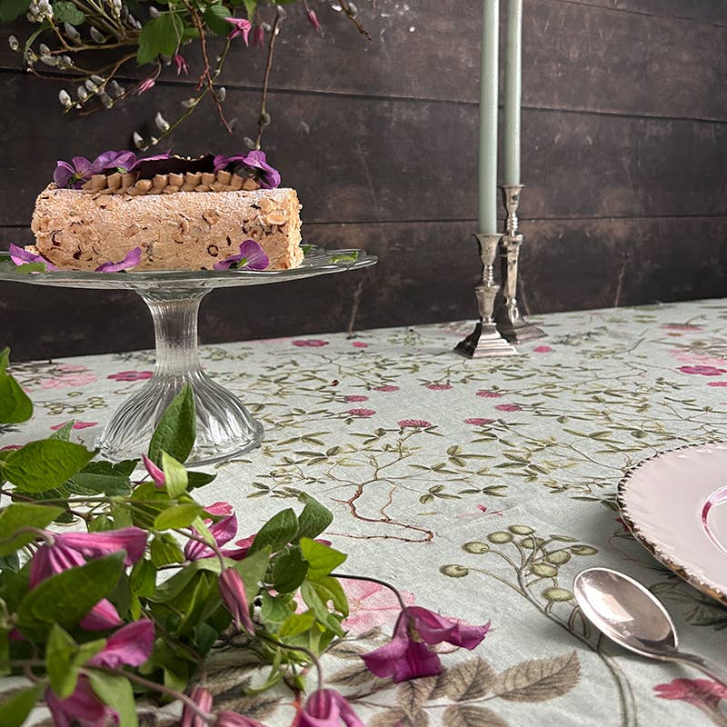 Ein Stück Kuchen mit violetten Blütenblättern liegt auf einem Glasständer inmitten der Tischdecke „Flora Danica Atlas“ von Koustrupco (350 x 145 cm), umgeben von grünen Kerzen in silbernen Haltern, einem Teller mit rosa Rand, einem Löffel und dekorativ angeordneten, blättrigen violetten Blumen.