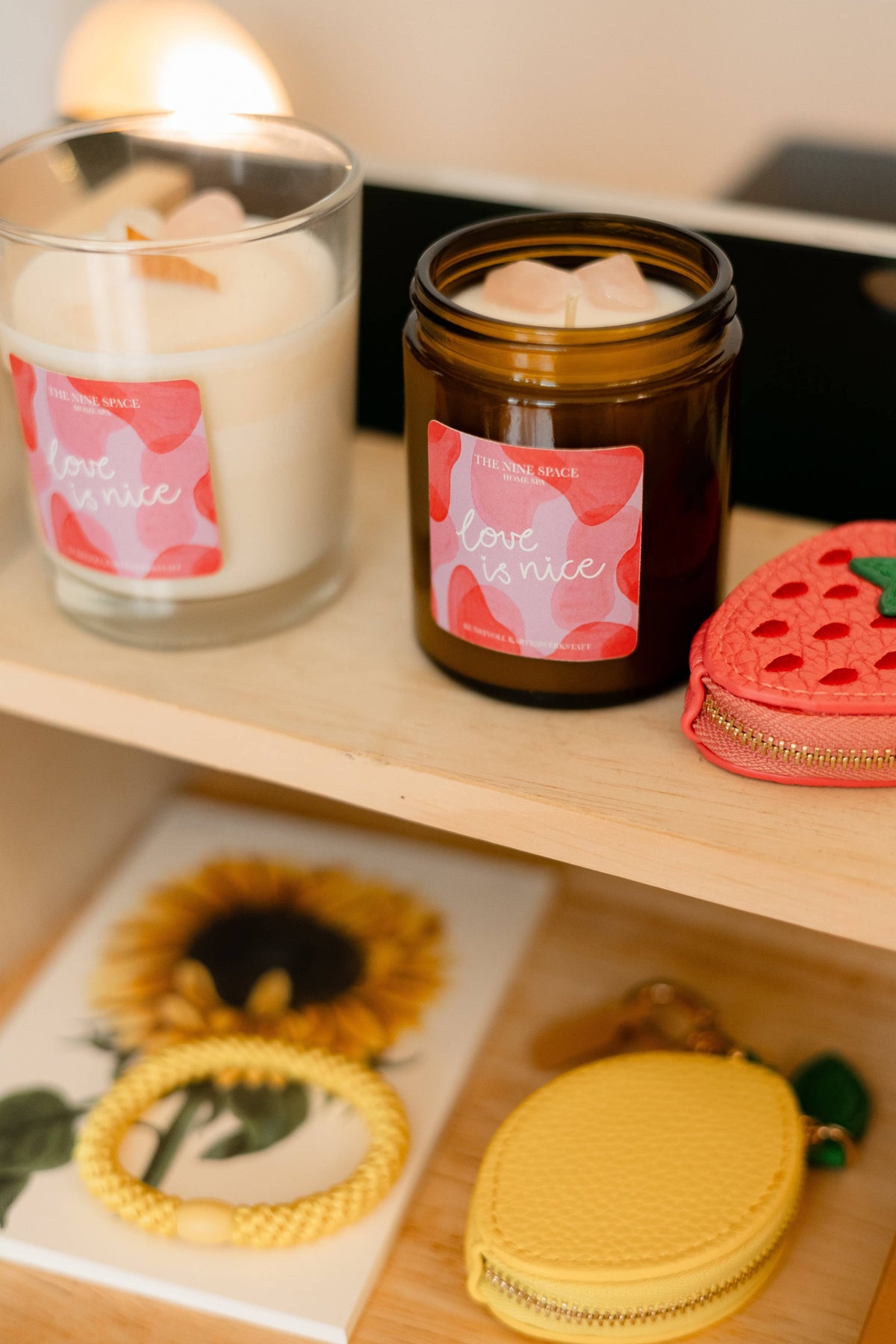 Auf einem Holzregal stehen zwei Kerzen mit der Aufschrift „love is nice“, ein roter Erdbeer-Beutel, ein Notizbuch mit Sonnenblumenmotiv sowie zwei gelbe Schlüsselanhänger aus Kunstleder in Apfel- und Birnenform von THE NINE SPACE.